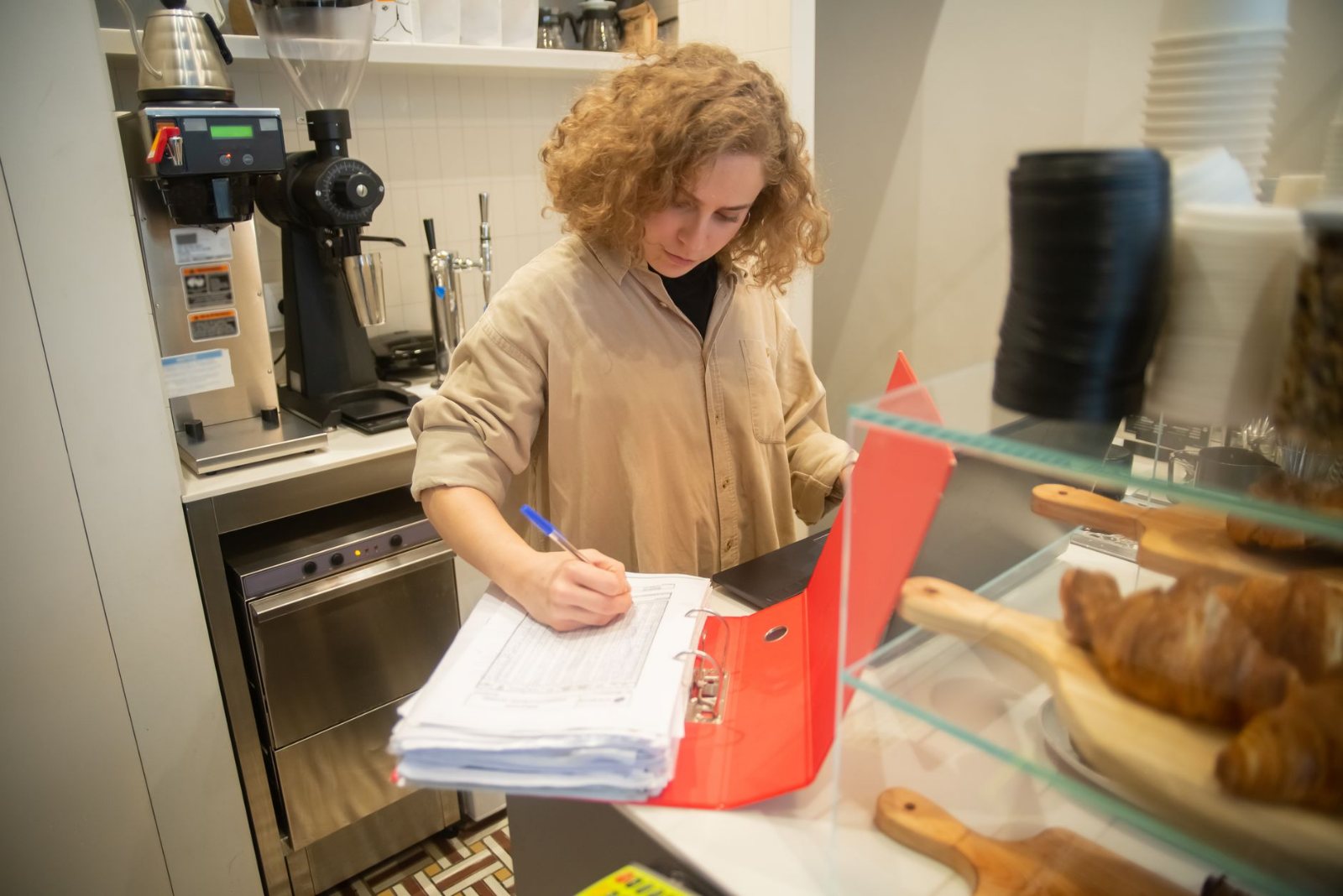 A small business owner writing in a labelled folder at her own shop counter, a receipt spike, a box of business cards and a closed laptop beside her, shelves softly out of focus behind.
